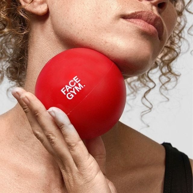 A close-up of a woman holding a vibrant red facial massage ball against her jawline, showcasing its smooth, glossy surface. The ball features the logo "FACE GYM" in bold white lettering. Curly hair frames her face, highlighting the minimalist aesthetic of her black top and the simplicity of the background.