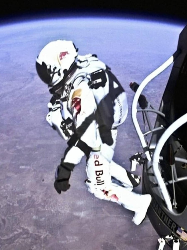 An astronaut in a white spacesuit adorned with the Red Bull logo steps out from a capsule against the backdrop of Earth's curvature, with a deep blue atmosphere transitioning to the vastness of space. The scene captures a moment of exploration and adventure, with the capsule's structure highlighting the extraordinary height.