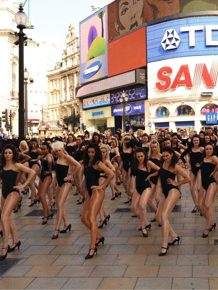 A vibrant scene in a bustling square filled with a large group of dancers, all adorned in sleek black leotards and high heels. The dancers stand in synchrony, showcasing their graceful forms against a backdrop of bright advertisements and historical architecture, bathed in warm sunlight.