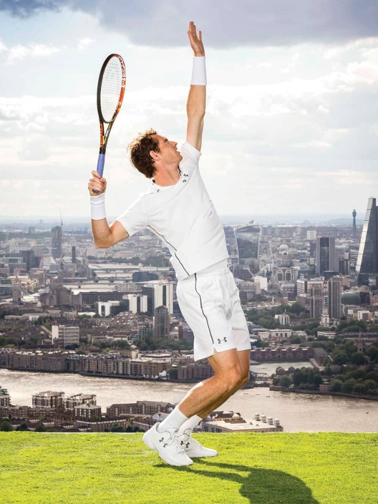 A male tennis player dressed in a white athletic outfit serves in a dynamic pose, lifted onto his toes. He holds a tennis racket high above his head, his eyes focused forward. In the background, a panoramic view of London unfolds, showcasing the Thames River and iconic skyscrapers under a partly cloudy sky.