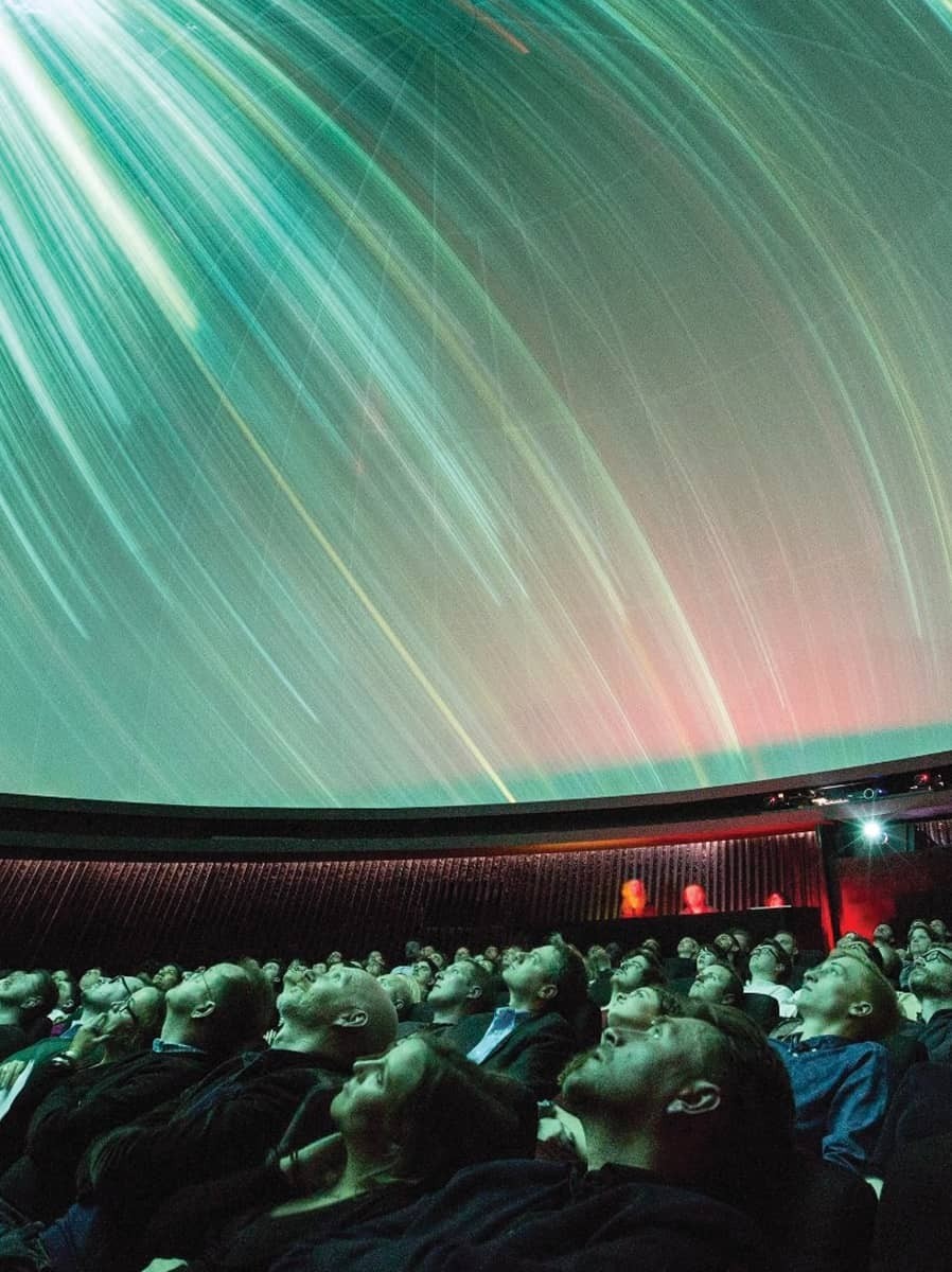 In a dimly lit planetarium, an audience gazes upwards at a mesmerizing display of swirling galaxy patterns projected onto a curved dome. Vivid streaks of light in hues of green and pink create an ethereal atmosphere, while spectators, captivated and still, are absorbed in the cosmic spectacle.