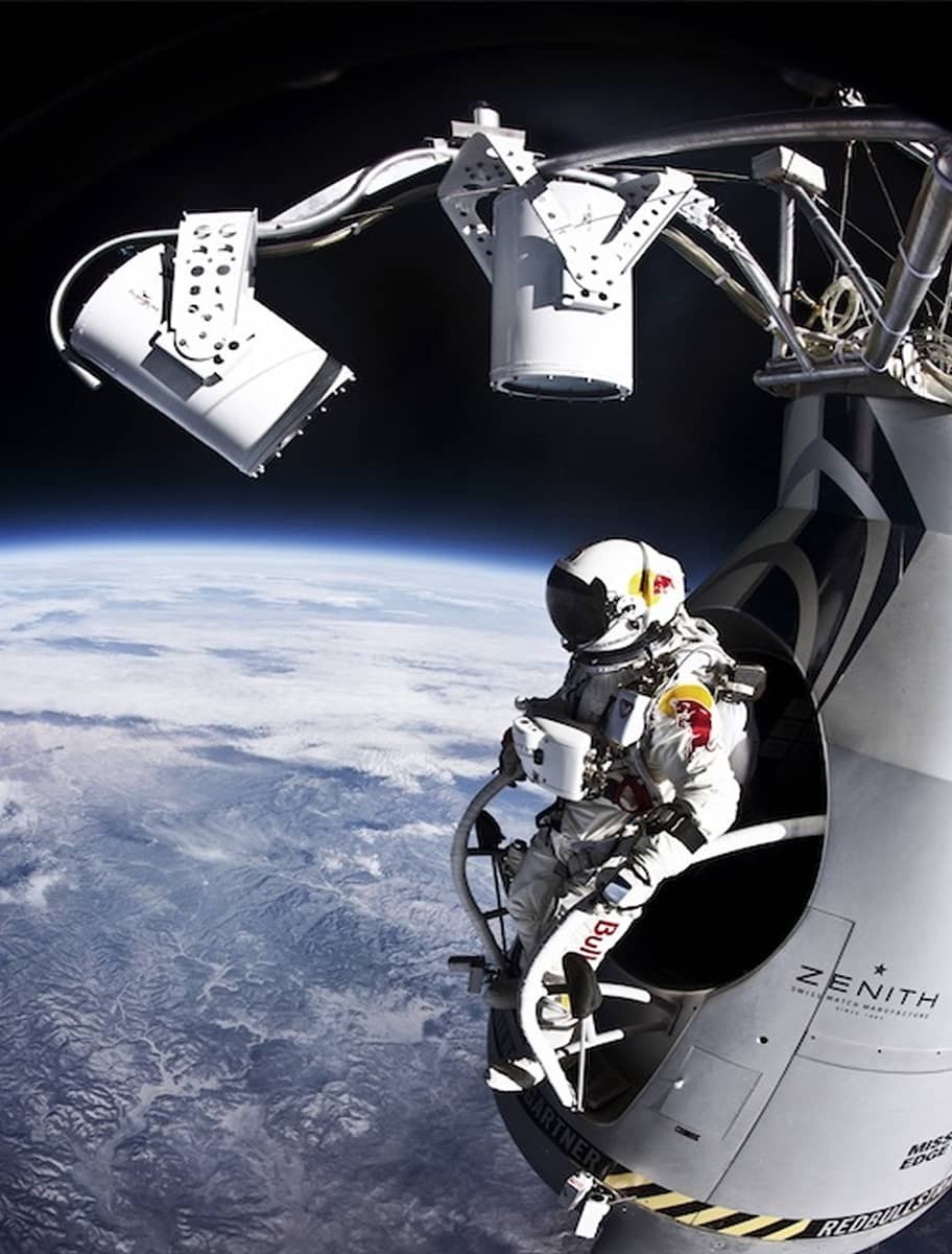 An astronaut in a sleek, white space suit, emblazoned with a vivid Red Bull logo, is seated on the edge of a floating capsule. Below is a breathtaking vista of Earth, with swirling white clouds and rugged terrains visible. The scene is illuminated by a soft, ethereal blue light, creating a serene yet exhilarating atmosphere.