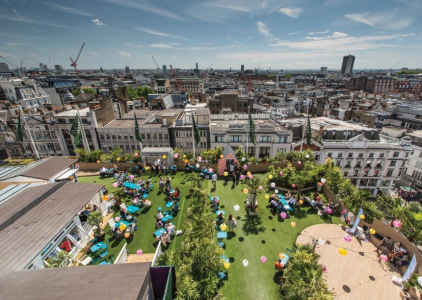 A vibrant rooftop garden is bustling with people celebrating a lively event in the heart of the city. Lush green grass contrasts with colourful balloons and chic teal seating. The skyline is dotted with historic rooftops and modern skyscrapers under a bright blue sky with wispy clouds.