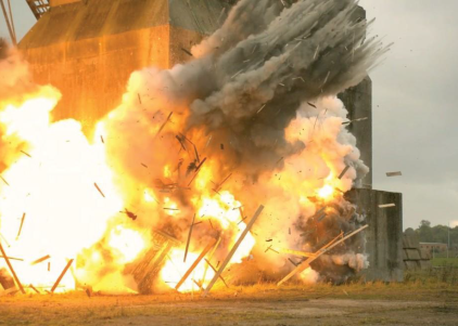 A dramatic explosion erupts from a weathered concrete structure, sending plumes of fire and billowing smoke into the air. Fragments of debris scatter widely, illuminated by the fierce orange glow of the blast, contrasting against a backdrop of overcast skies.