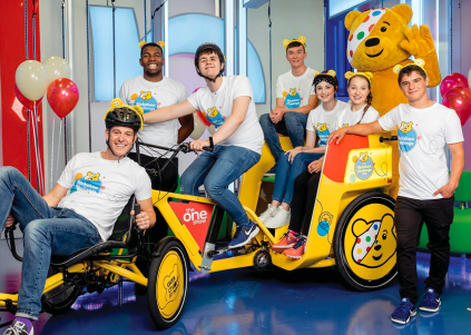 A lively group of six young adults, all wearing cheerful white t-shirts featuring the iconic Pudsey Bear, gather around a bright yellow pedicab. The pedicab, adorned with the BBC Children in Need logo, stands against a playful backdrop of colourful balloons and studio lights, creating a vibrant atmosphere.