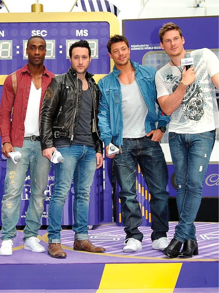 Four men pose against a vibrant backdrop showcasing a purple and yellow colour scheme. The first, in a red checked shirt, stands confidently beside a man in a black leather jacket. The third sports a denim jacket over a white shirt, while the fourth wears a graphic tee with denim jeans. The scene exudes a lively, modern vibe.