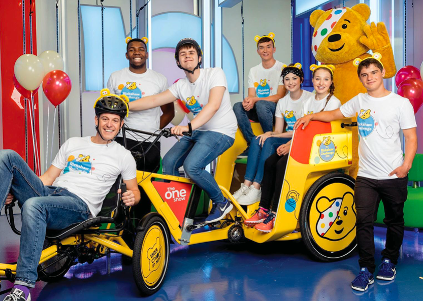 A lively group of six young adults, all wearing cheerful white t-shirts featuring the iconic Pudsey Bear, gather around a bright yellow pedicab. The pedicab, adorned with the BBC Children in Need logo, stands against a playful backdrop of colourful balloons and studio lights, creating a vibrant atmosphere.