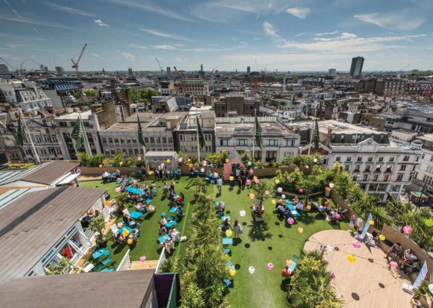 A vibrant rooftop garden is bustling with people celebrating a lively event in the heart of the city. Lush green grass contrasts with colourful balloons and chic teal seating. The skyline is dotted with historic rooftops and modern skyscrapers under a bright blue sky with wispy clouds.