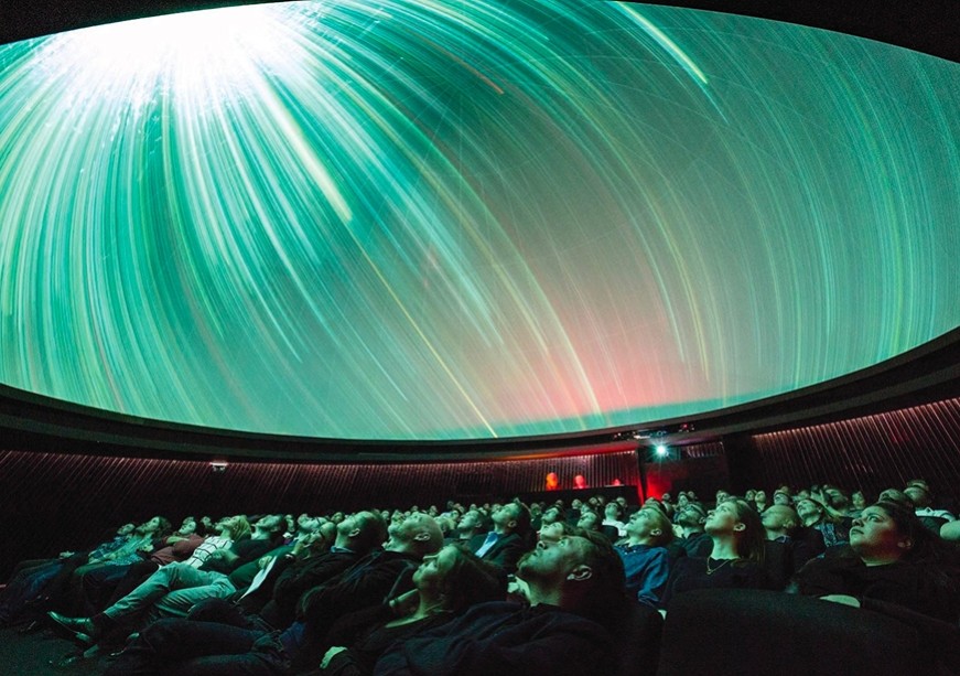 Inside a planetarium, a diverse audience reclines on cushioned seats, mesmerised by a vibrant, swirling green and blue light display on the domed ceiling. The room is softly illuminated, creating an immersive atmosphere that evokes a cosmic wonder.