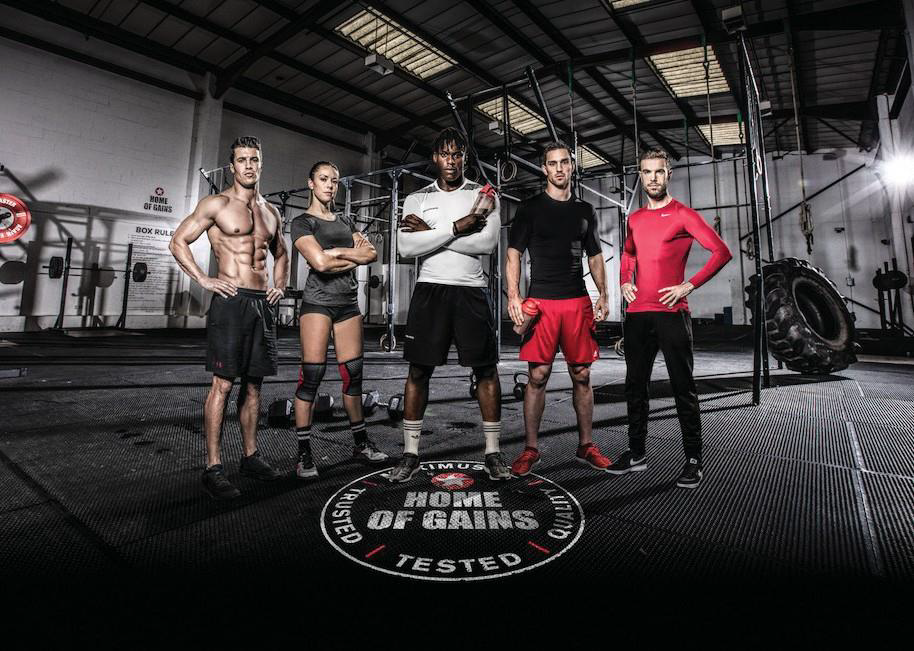A diverse group of five athletes stands confidently in a well-lit gym. The men and woman showcase a range of athletic attire, including fitted shorts and compression shirts. The industrial backdrop features gym equipment, creating an atmosphere of determination and strength, with the 'Home of Gains' logo prominent on the floor.