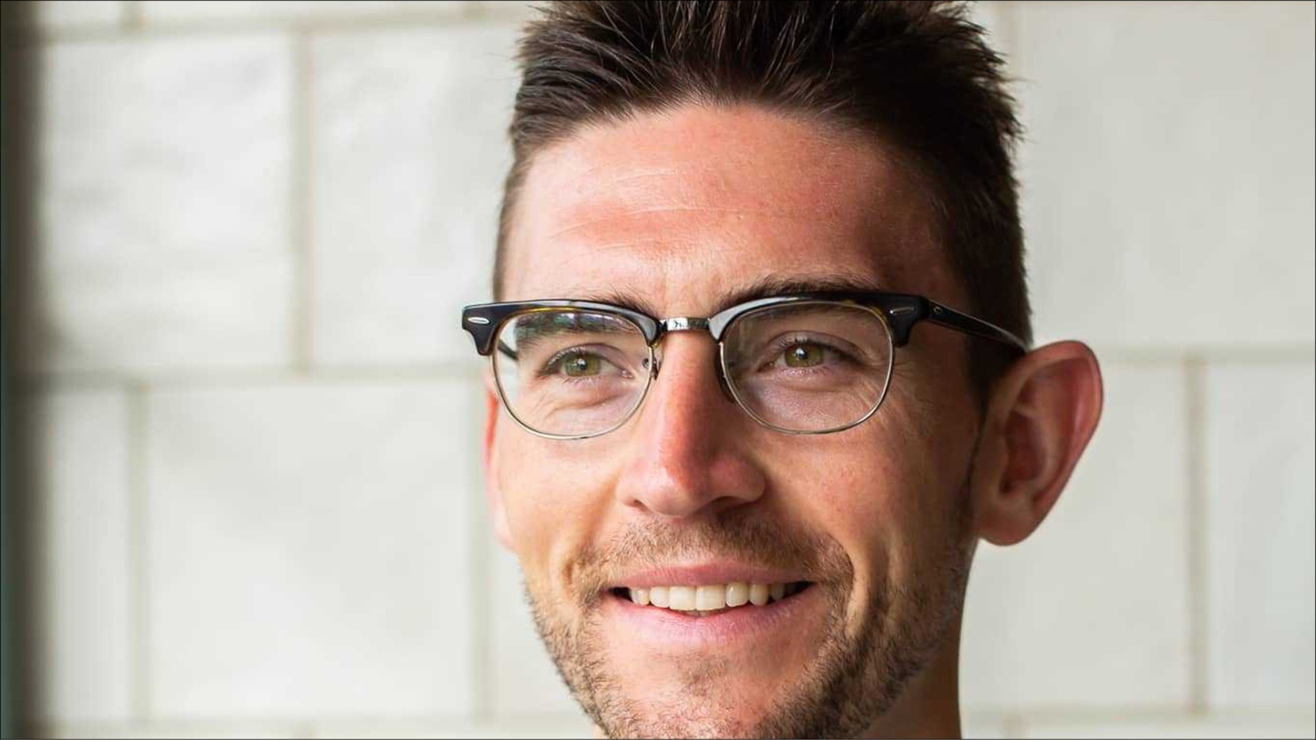 A young man with short, dark hair and a neatly trimmed beard, wearing stylish black glasses. He exudes an air of confidence and approachability, smiling gently against a backdrop of light grey tiled walls that enhance the modern, fresh atmosphere.