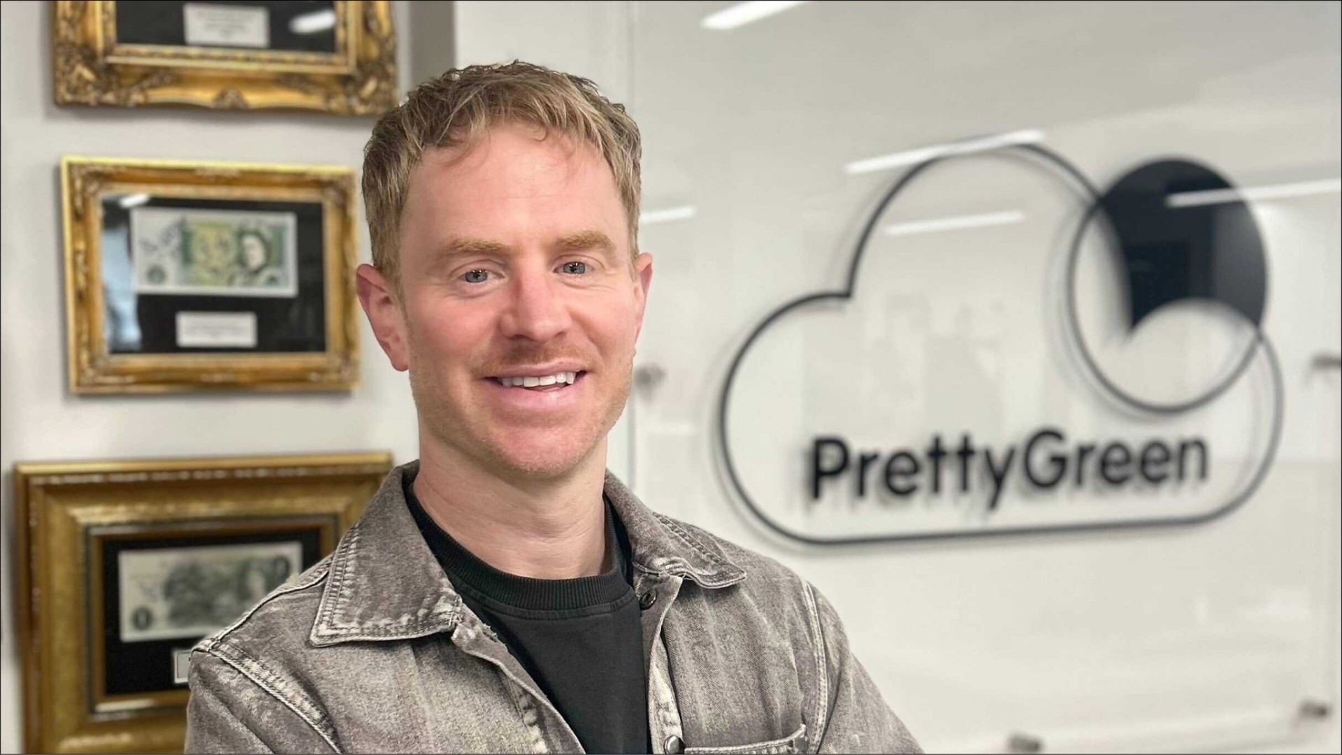 A smiling man with short, wavy blonde hair stands in an office environment. He wears a light grey denim jacket over a black shirt. Behind him, a stylish logo for 'PrettyGreen' is displayed on a glass wall, accompanied by ornate picture frames showcasing dollar bills.