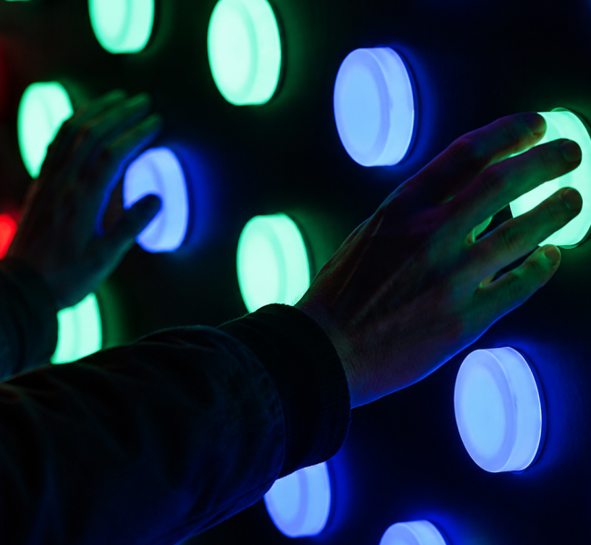 Two hands gently pressing against a wall illuminated by a vibrant display of circular lights in various colours: bright green, blue, and red. The hands are silhouetted against the dark background, creating a striking contrast that enhances the luminescent glow of the buttons.