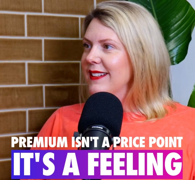 A woman with long, wavy blonde hair speaks into a black microphone against a backdrop of glossy brown tiles interspersed with large green leaves. She wears a vibrant coral top, creating a lively contrast with the earthy tones behind her. Bold text at the bottom emphasizes a message about the emotional essence of premium experiences.