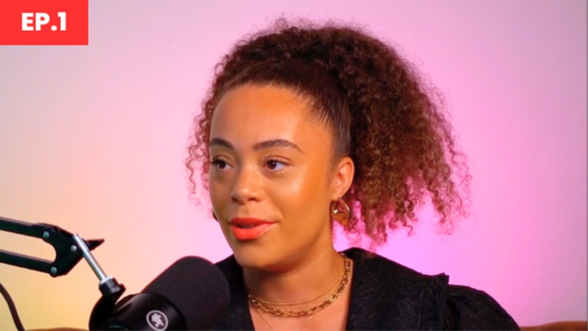 A young woman with curly hair styled in a high ponytail sits with poise in a dimly lit space. She wears a black, textured top and delicate gold jewellery. A microphone is positioned in front of her, while a warm, gradient backdrop enhances the intimate atmosphere.