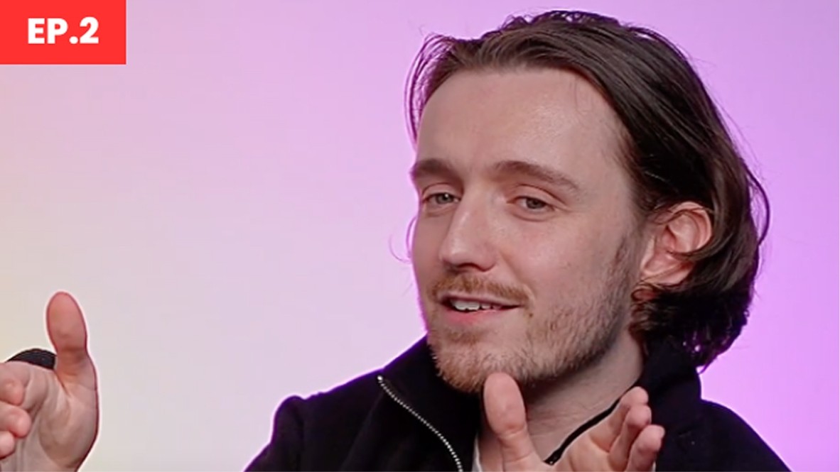 A man with tousled light brown hair gestures animatedly with both hands against a softly lit background transitioning from lavender to pale pink, enhancing the overall warmth of the scene. He wears a dark zip-up jacket, contributing a casual yet stylish aesthetic. A red graphic in the upper corner reads 'EP.2', further framing the image.