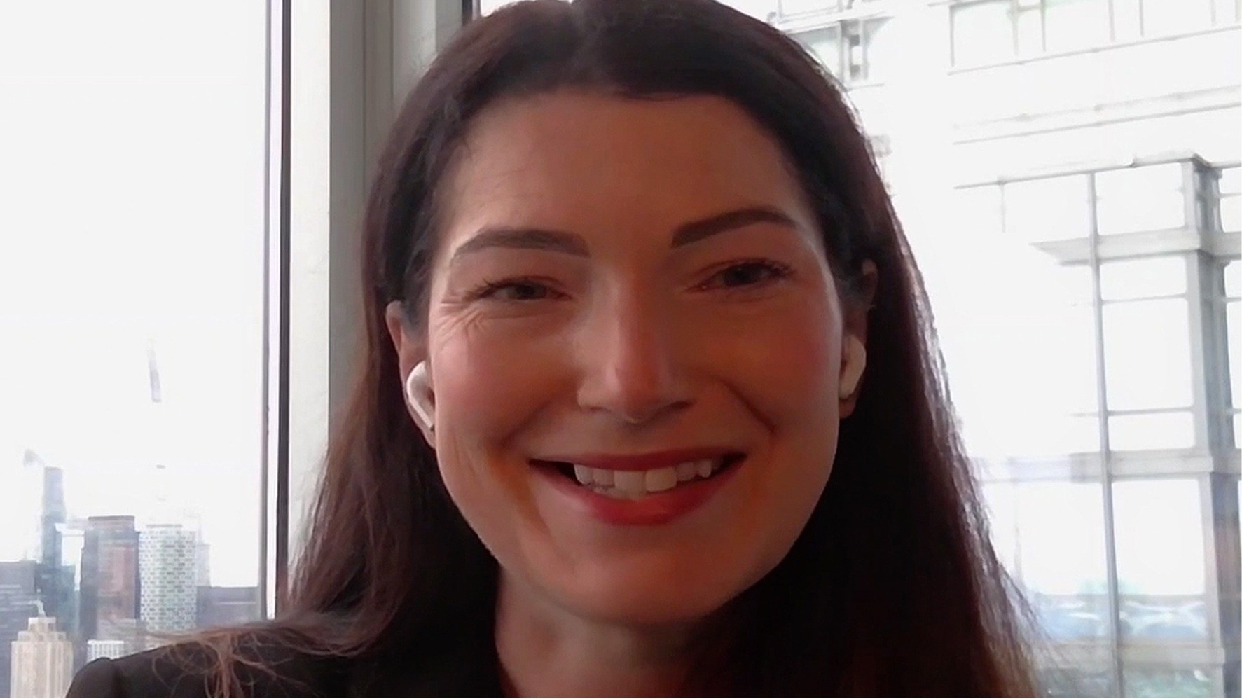 A woman with long dark hair framed by soft waves smiles warmly while wearing white earbuds. She is positioned in front of a large window, which lets in natural light that gently illuminates her features. The blurred cityscape in the background hints at a bustling urban environment.