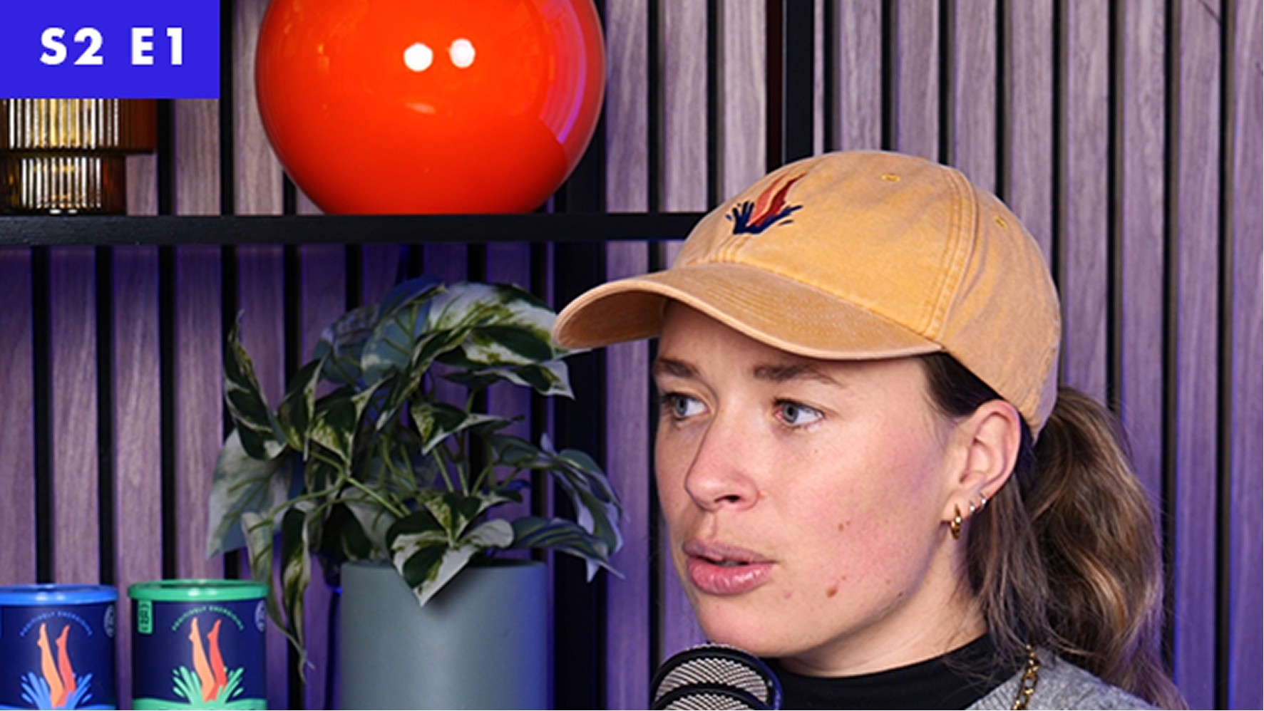 A woman with long brown hair wearing a soft yellow cap, stands beside a stylish arrangement of vibrant objects. Behind her, a plant flourishes with lush green leaves, while a round orange lamp adds a pop of colour. The setting features sleek shelves in a chic, contemporary style, enhancing the modern aesthetic.