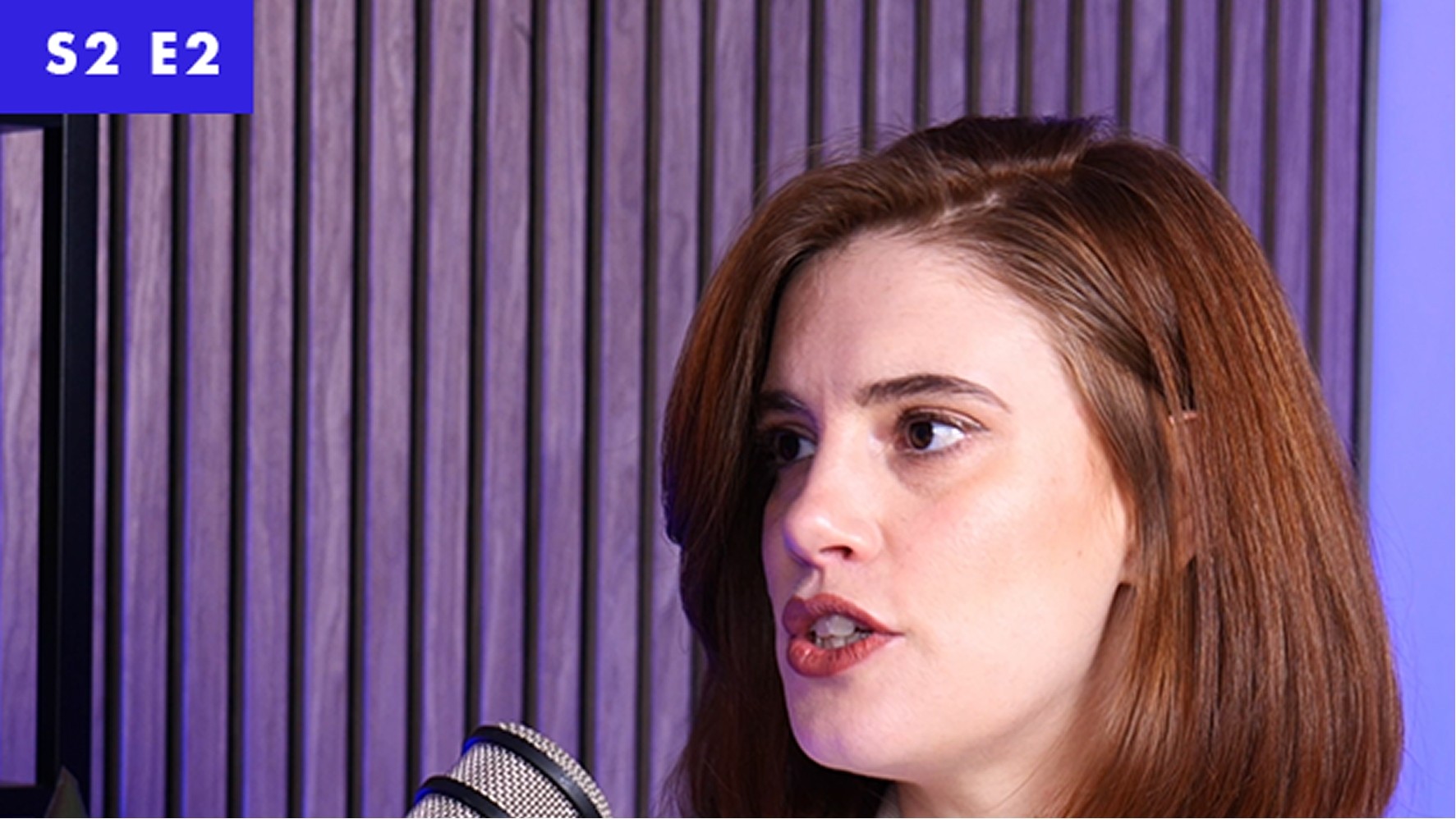 A woman with long, auburn hair speaks passionately into a microphone, framed by a modern studio environment. The backdrop features textured wooden panels lit by soft purple lighting, enhancing the contemporary aesthetic. In the corner, a bold purple graphic displays "S2 E2".