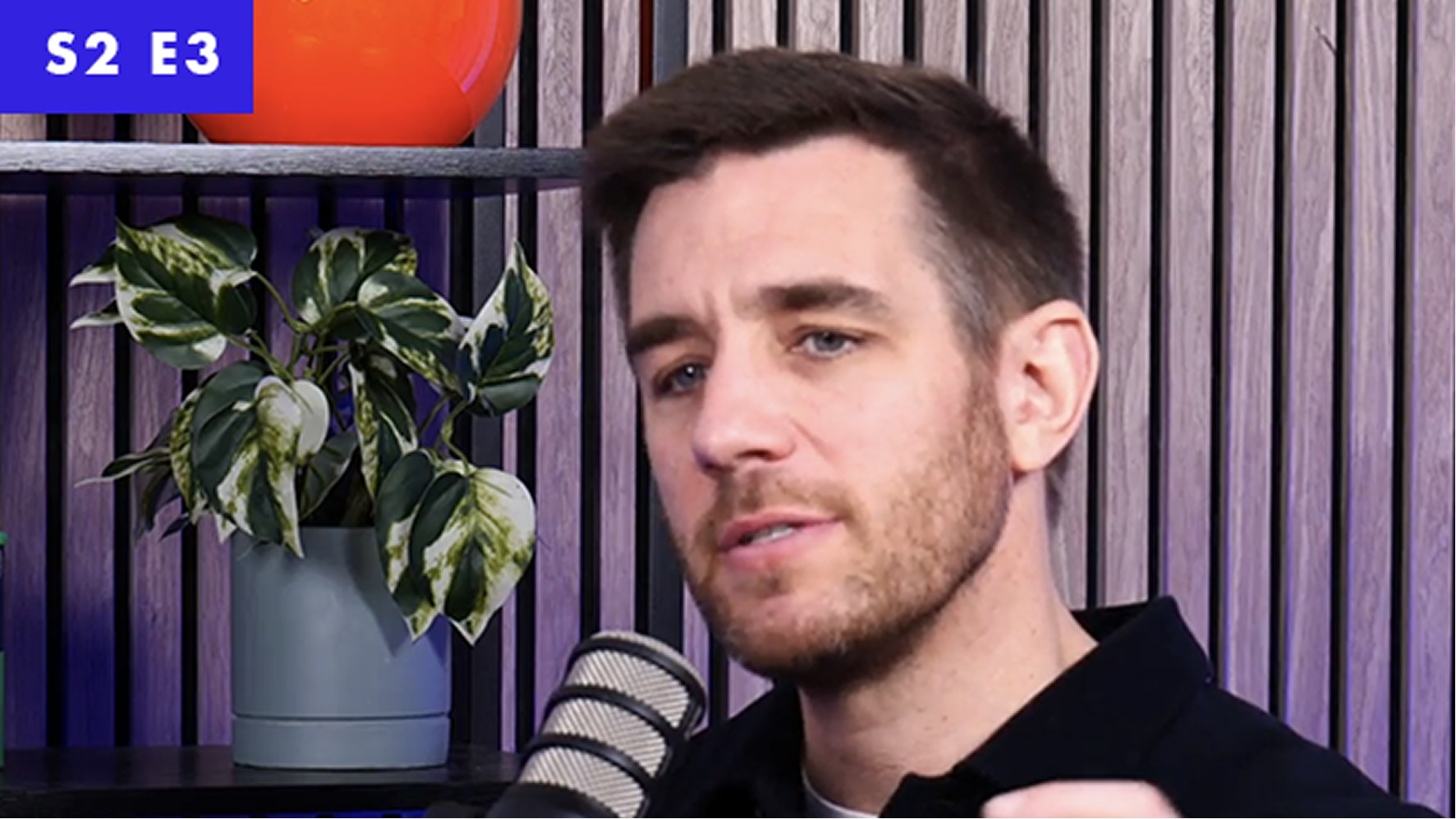 A man with short brown hair, dressed in a black shirt, gestures thoughtfully during a discussion. Behind him, a minimalist shelf holds a vibrant orange vase and a leafy green plant in a soft grey pot. The background features a modern wooden wall with vertical slats, creating a contemporary atmosphere.