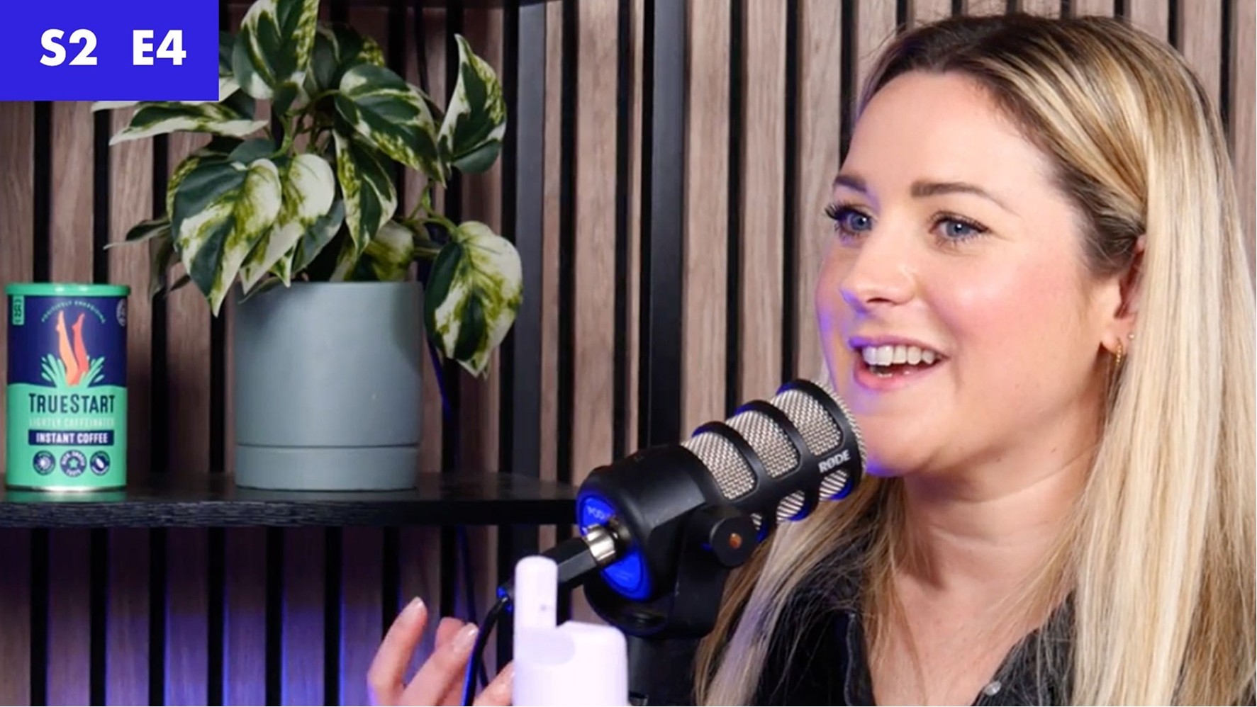 A woman with blonde hair, styled in loose waves, sits in front of a sleek black microphone, engaged in conversation. Behind her, a vibrant green plant thrives in a minimalist grey pot, complemented by a trendy energy drink can. The warm wooden backdrop adds depth, while soft lighting enhances the inviting atmosphere.