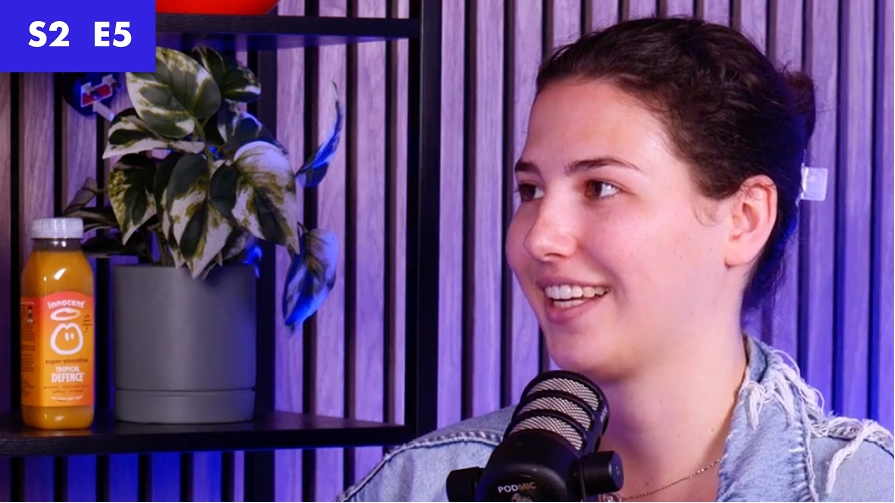 A young woman with dark hair sits in front of a stylish microphone, her denim jacket contrasting with the vibrant blue-lit background. A lush green plant and a bottle of orange drink rest on a black shelf, adding warmth to the modern interior.