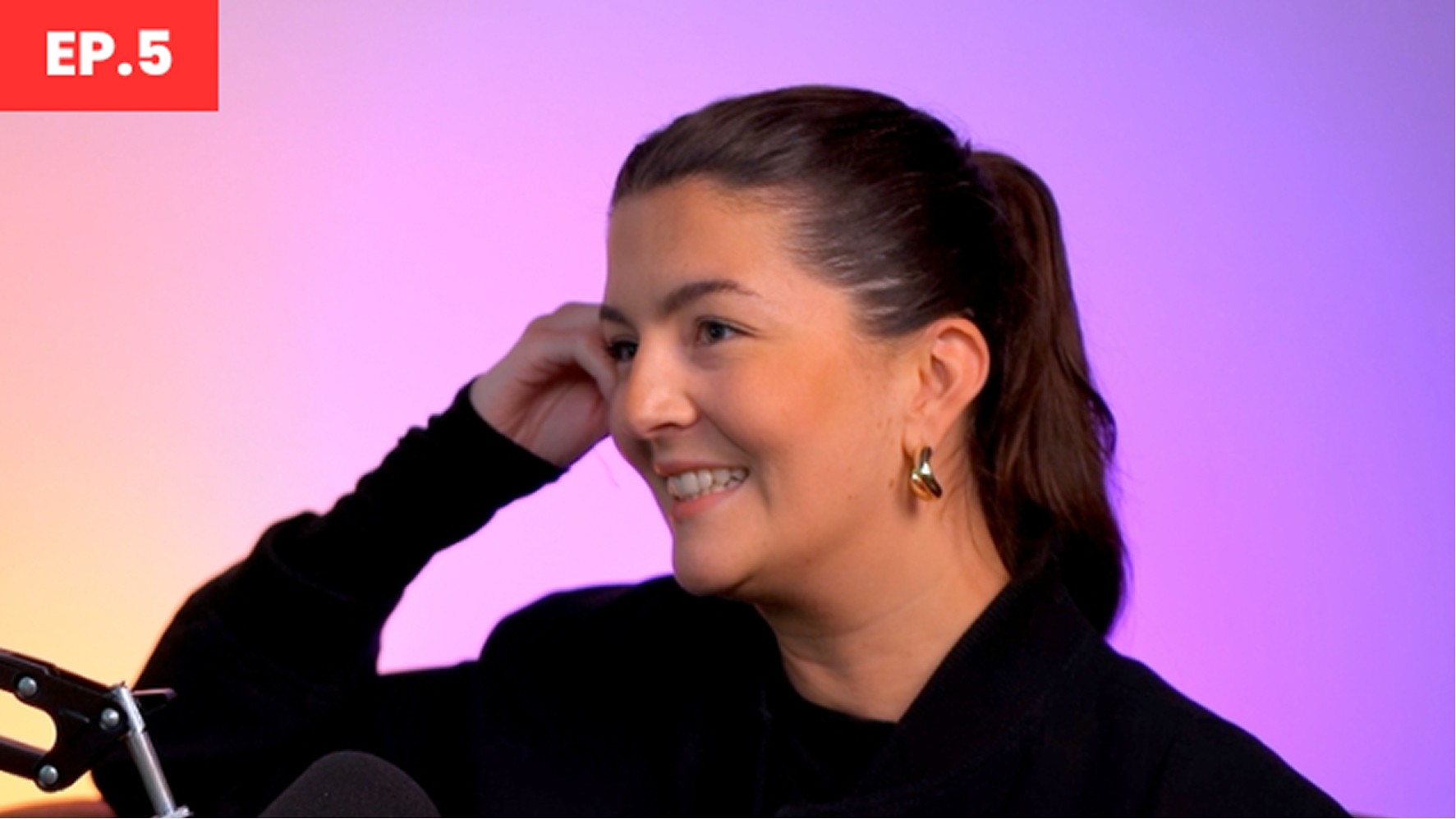 A woman with dark hair tied back in a sleek ponytail touches her head lightly, wearing an elegant black outfit complemented by gold hoop earrings. The background transitions from soft lavender to deep purple, creating a calming yet vibrant atmosphere, while a microphone is positioned in front of her.