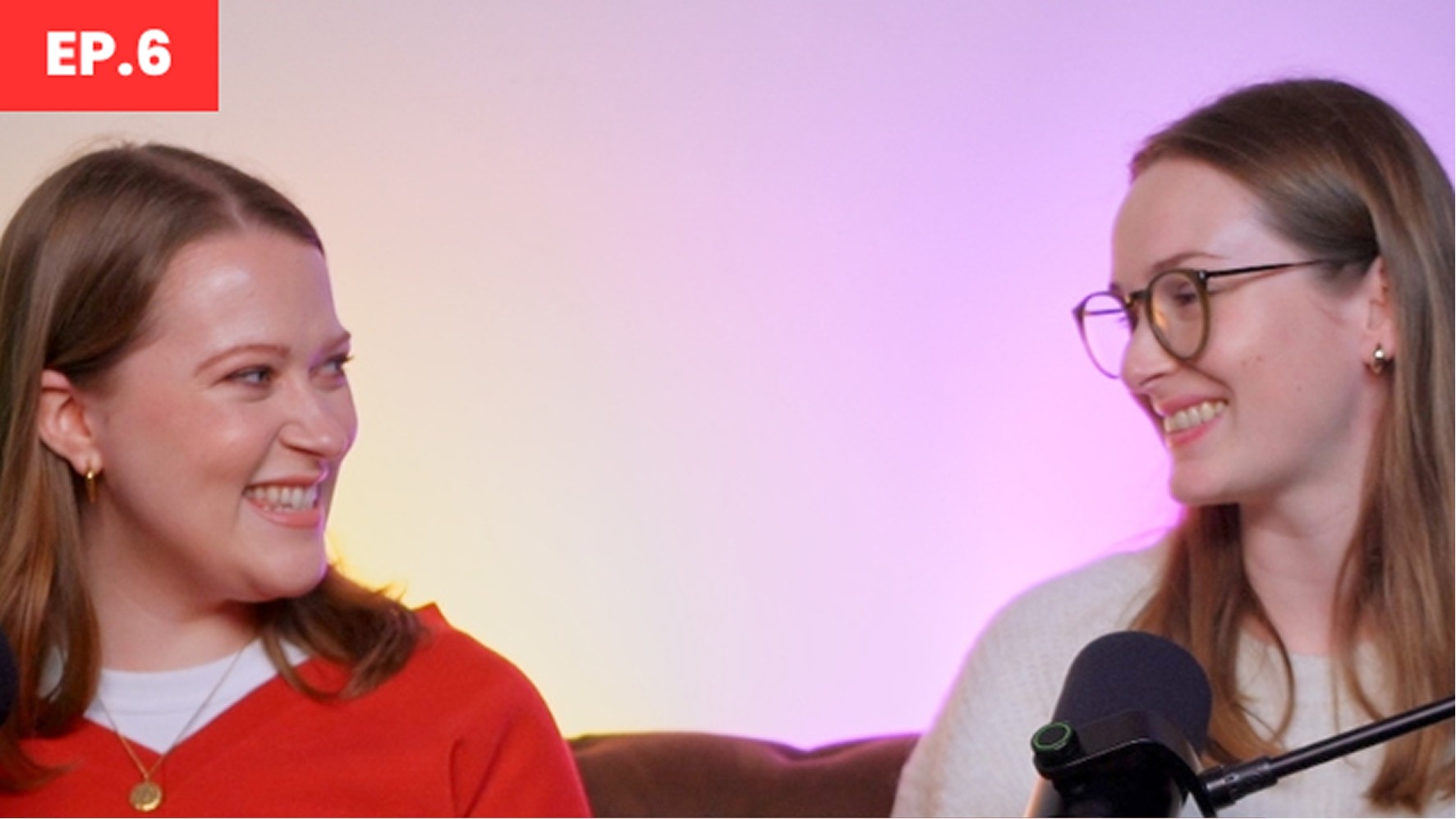 Two women sit side by side in a cozy podcast setting adorned with soft, warm lighting that transitions from yellow to purple in the background. The woman on the left, dressed in a vibrant red sweater, exudes a refined style with a delicate necklace, while the second woman, in a light-coloured knit sweater, sports stylish glasses. A microphone stands before them, inviting engaging conversation.