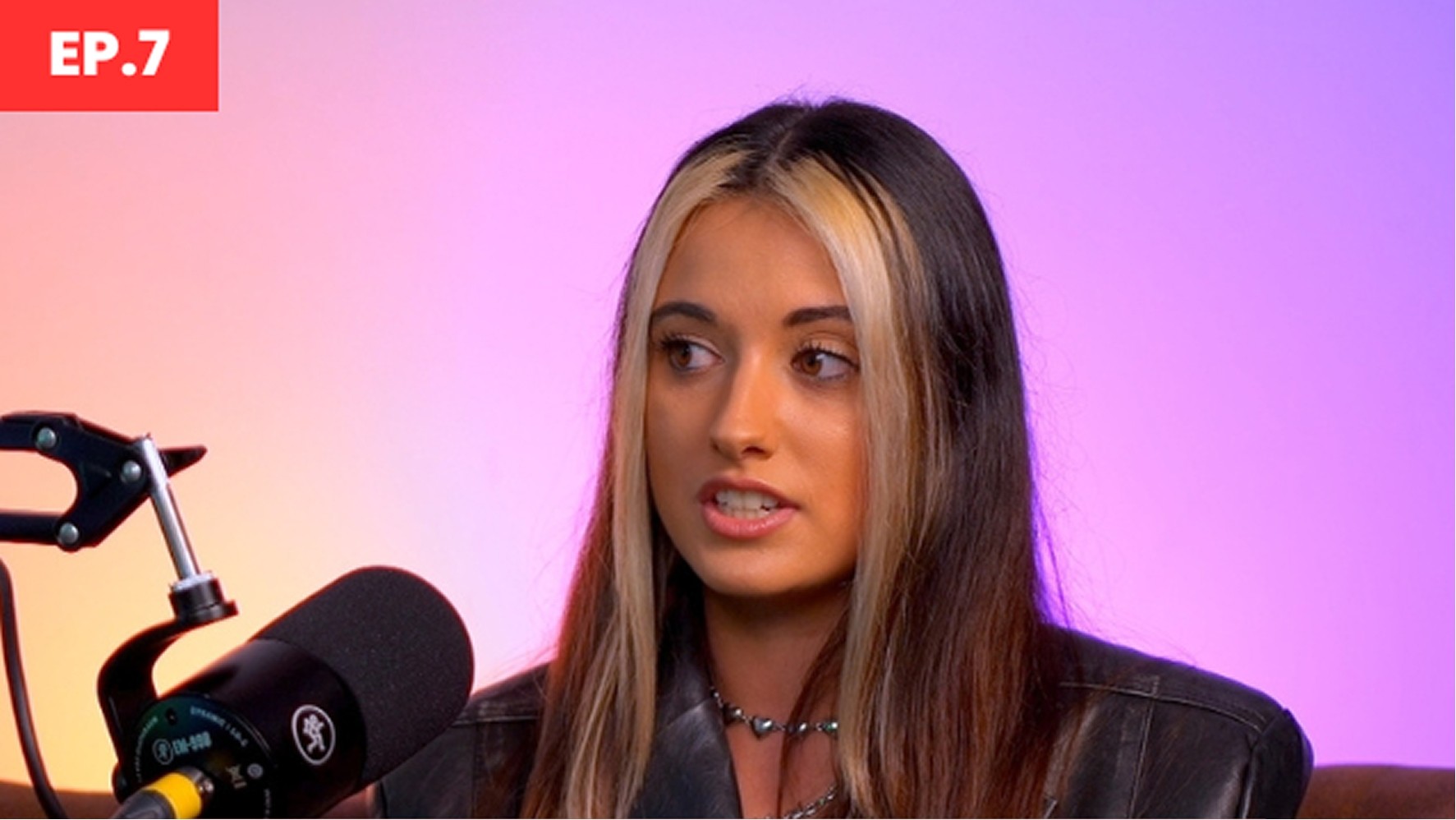 A young woman with long, flowing hair featuring blonde highlights, wears a leather jacket over a black top, seated in a modern studio. A microphone is positioned close to her, while a vibrant gradient backdrop transitions from purple to pink, creating a lively atmosphere.