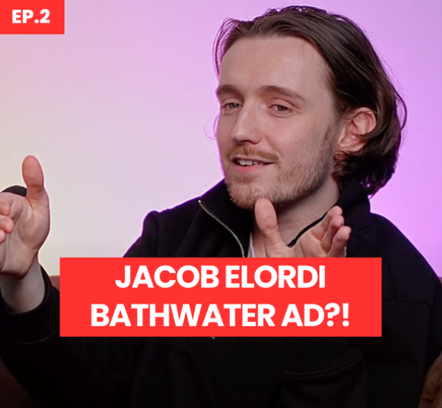 A young man with medium-length brown hair gestures animatedly while discussing a topic. He wears a stylish black jacket against a softly lit background, featuring a gradient of warm hues. The image prominently showcases bold text stating, 'JACOB ELORDI BATHWATER AD?!', adding a striking contrast.