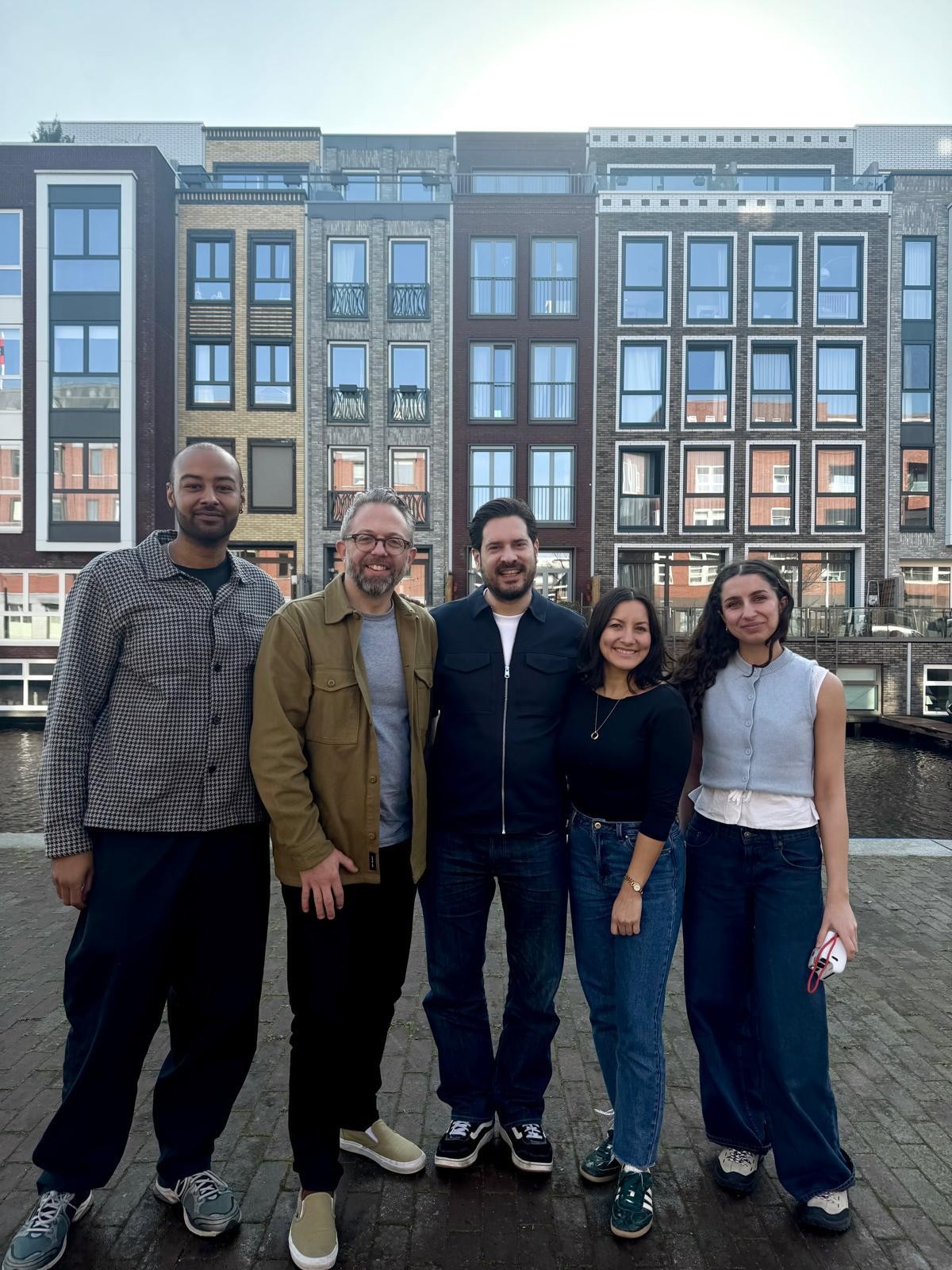 A group of five individuals stands together on a cobblestone walkway beside a canal. Behind them looms an array of modern and historic brick buildings with large, rectangular windows. The sunlight casts a soft glow, illuminating their relaxed attire, creating a lively urban atmosphere.