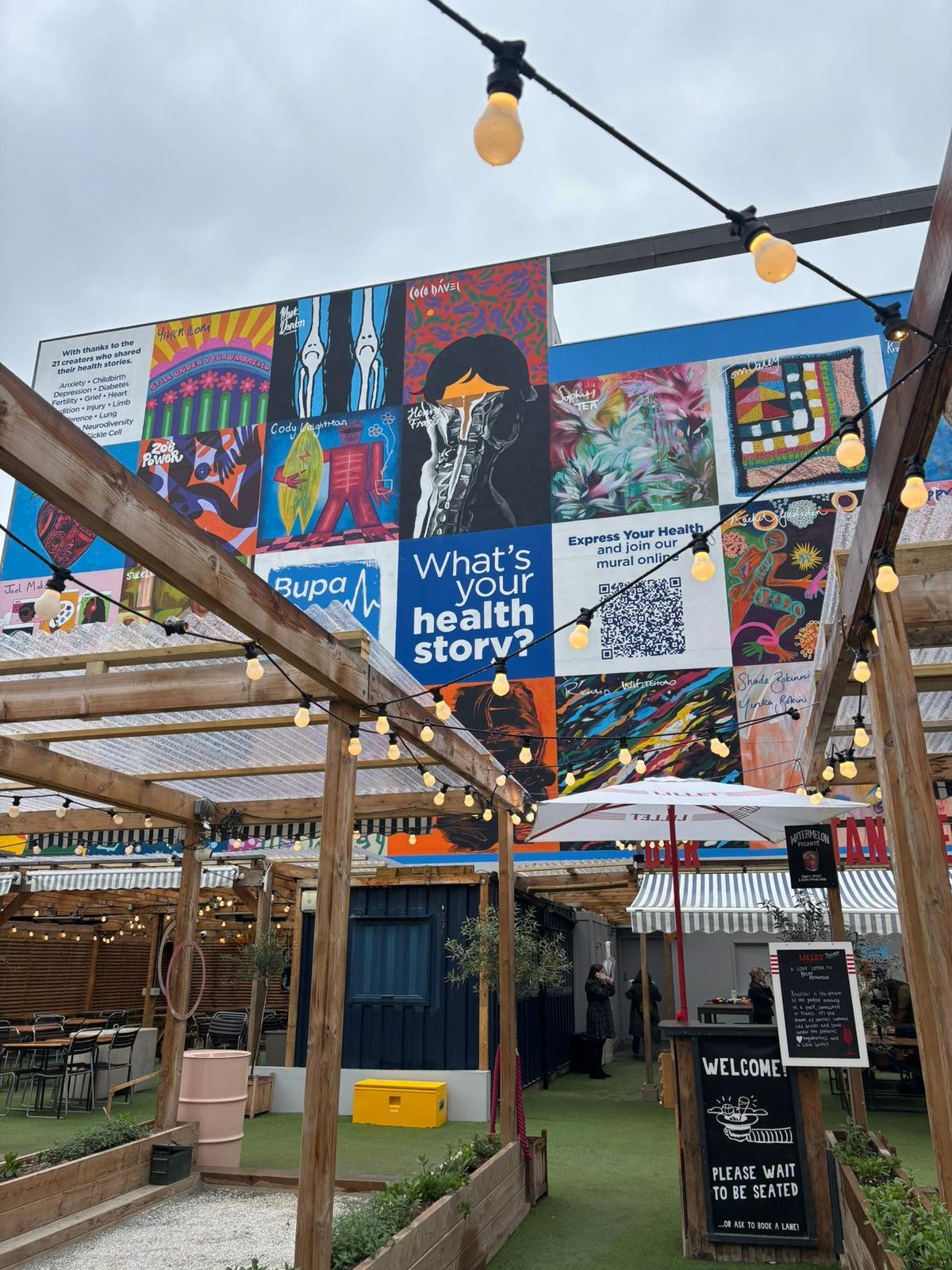 An outdoor venue adorned with warm string lights, highlighted by a vibrant mural displaying colourful graphics and text that asks, "What's your health story?" The atmosphere is lively and inviting, with rustic wooden structures framing the grassy area filled with scattered seating.