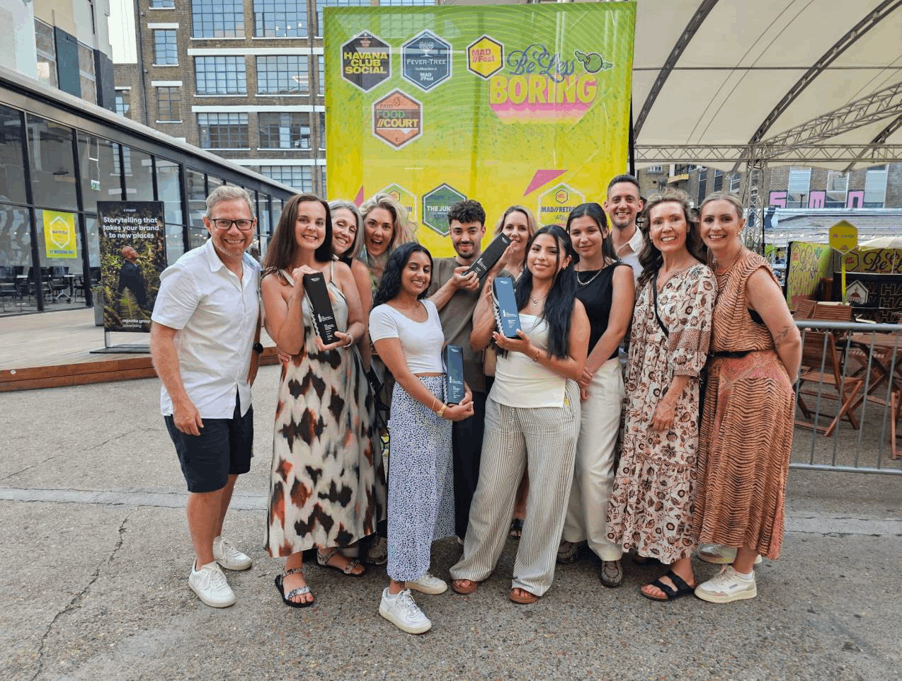 A cheerful group of eleven individuals poses for a photo in a vibrant outdoor setting. They are dressed in a mix of fashionable summer outfits, showcasing flowing dresses and stylish shorts. Behind them, a bright neon banner reads ‘Not Boring’, adding to the lively atmosphere.
