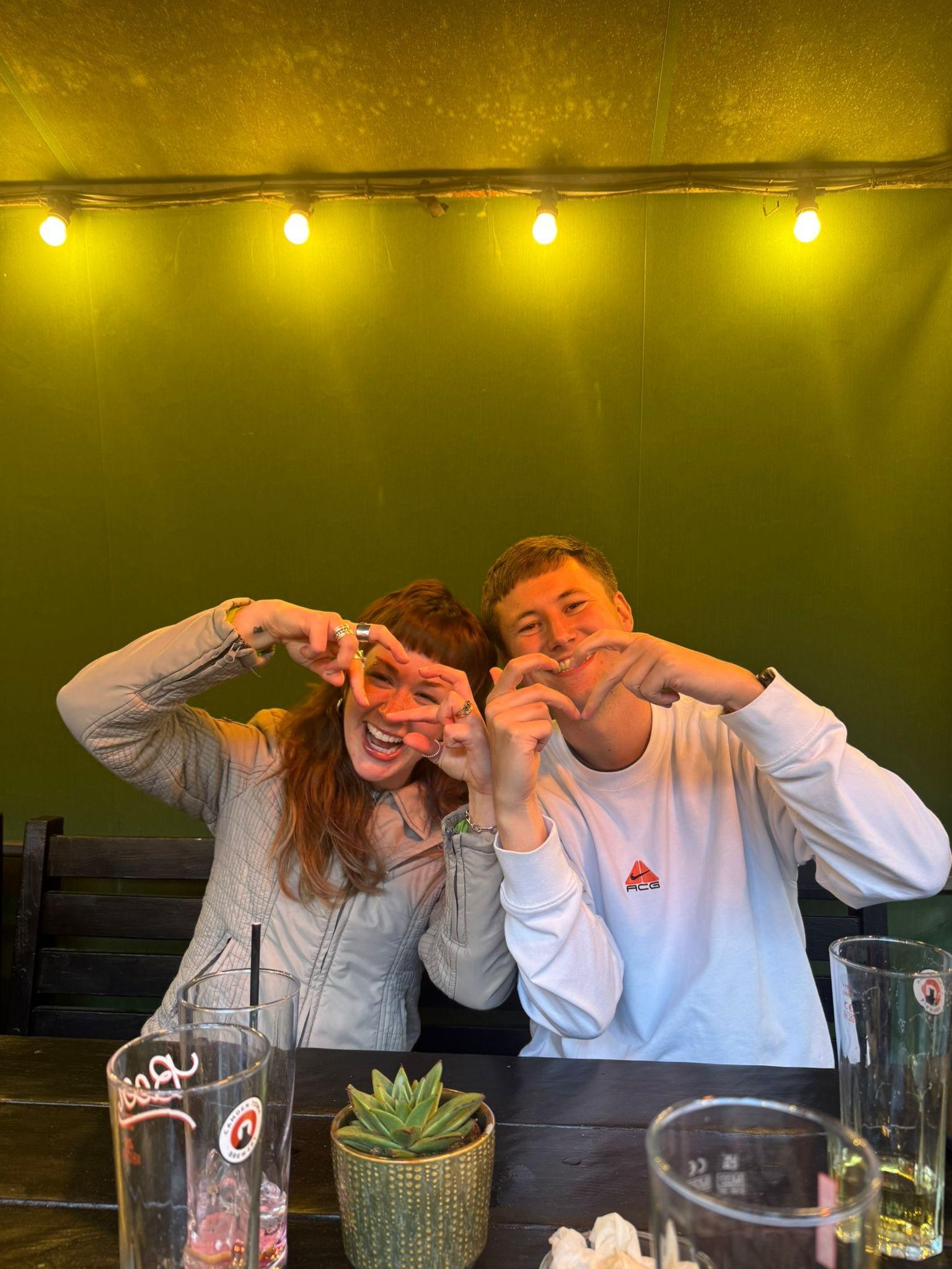 Against a backdrop of glowing yellow fairy lights and lush green walls, a woman with flowing auburn hair playfully forms a heart shape with her hands next to a man in a white sweatshirt. Their joyful expressions reflect a lively moment in a vibrant social setting.