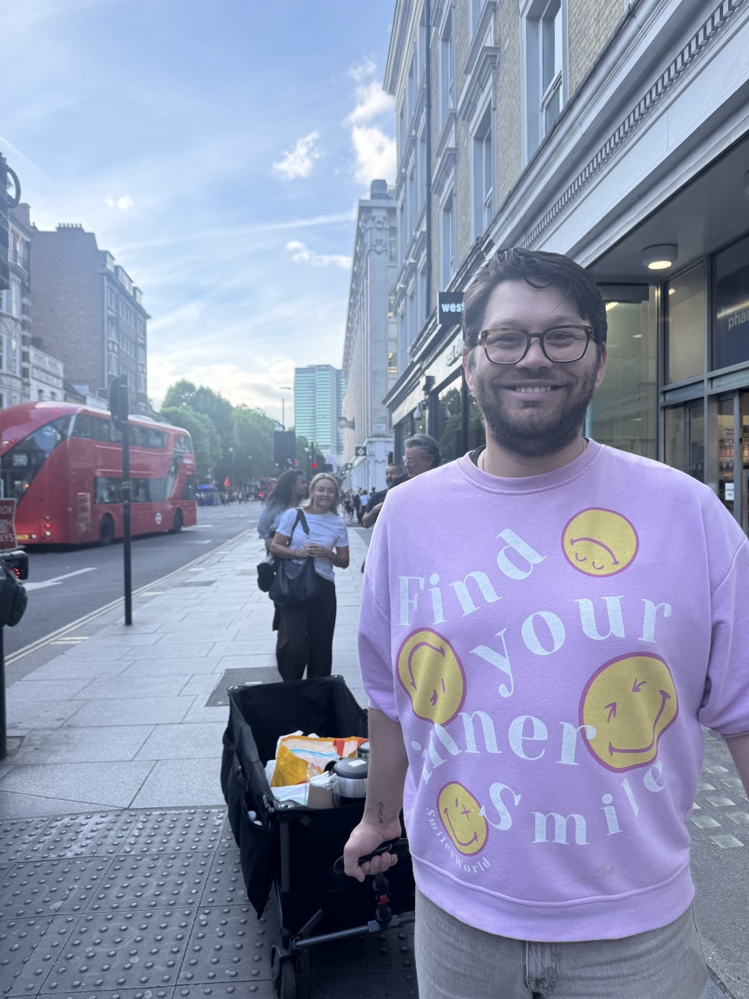 A young man stands confidently on a bustling city street, wearing a playful pink sweatshirt adorned with cheerful smiley faces. Behind him, a cart filled with groceries contrasts with the vibrant red double-decker buses and historic architecture lining the road. Sunlight bathes the scene, enhancing the lively urban atmosphere.