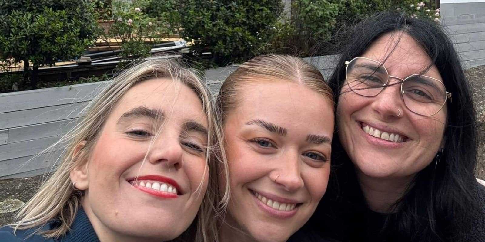 Three women pose closely together for a cheerful selfie, framed by lush greenery behind them. The first woman on the left has long, wavy blonde hair and bold red lips, wearing a navy jacket. The centre woman, with smooth fair hair, beams brightly. The woman on the right, in black attire with glasses, has flowing dark hair. Their warm smiles radiate joy.
