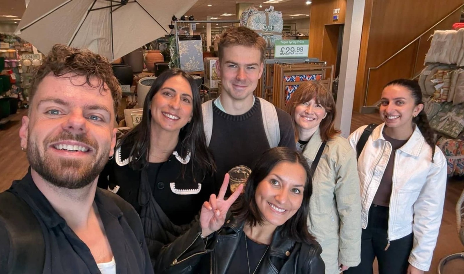 A group of six friends gathers in a well-lit store featuring home décor. The vibrant scene showcases a cheerful atmosphere, with textiles and decorative items in the background. Sunlight filters through a large window, illuminating their camaraderie as they pose happily for a selfie.
