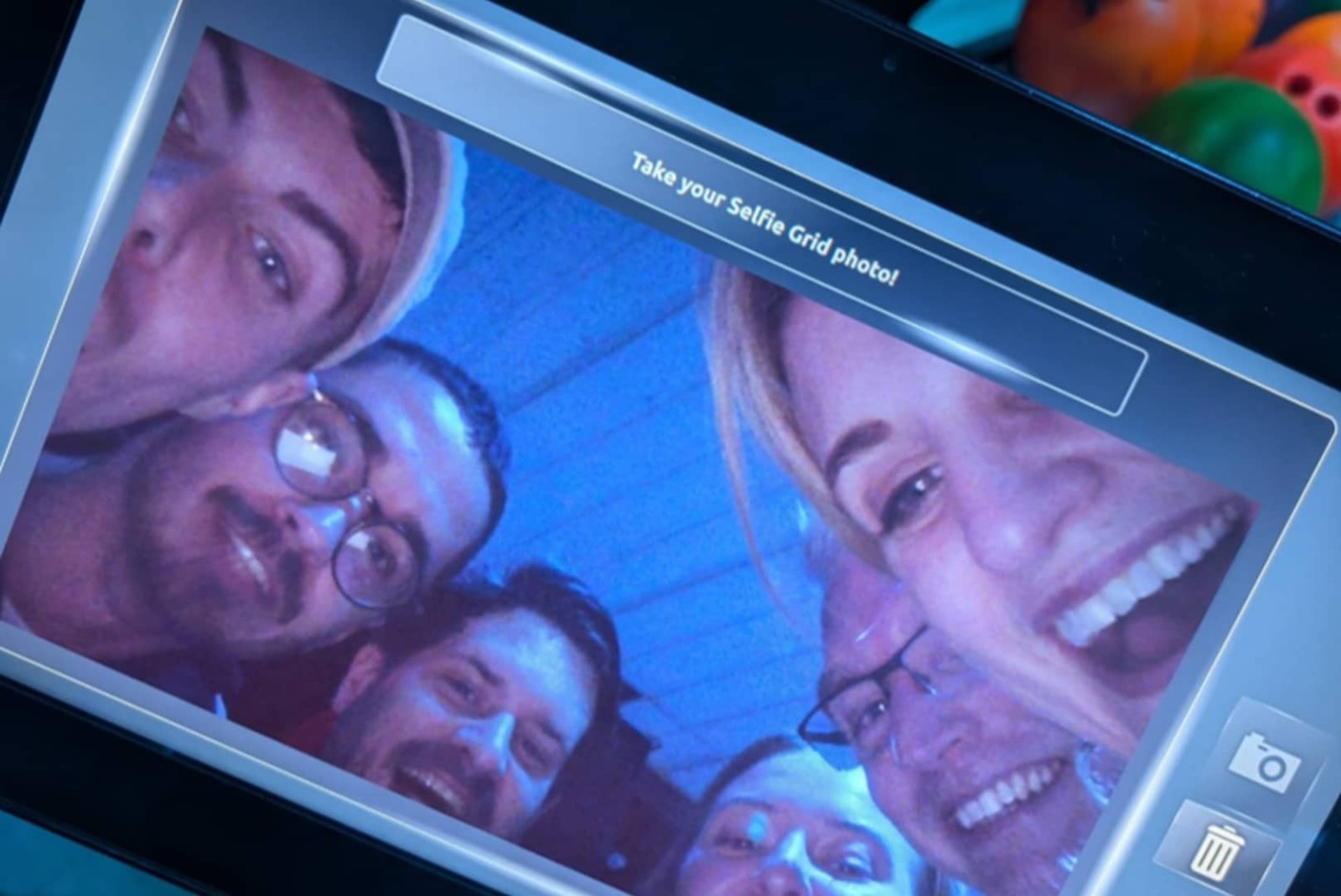 A close-up shot of a group of five people smiling and posing for a selfie, captured on a digital screen. The background is illuminated in soft blue light, creating a cheerful atmosphere. Each person shows a distinct style, with varying hairstyles and accessories, enhancing the lively group dynamic.