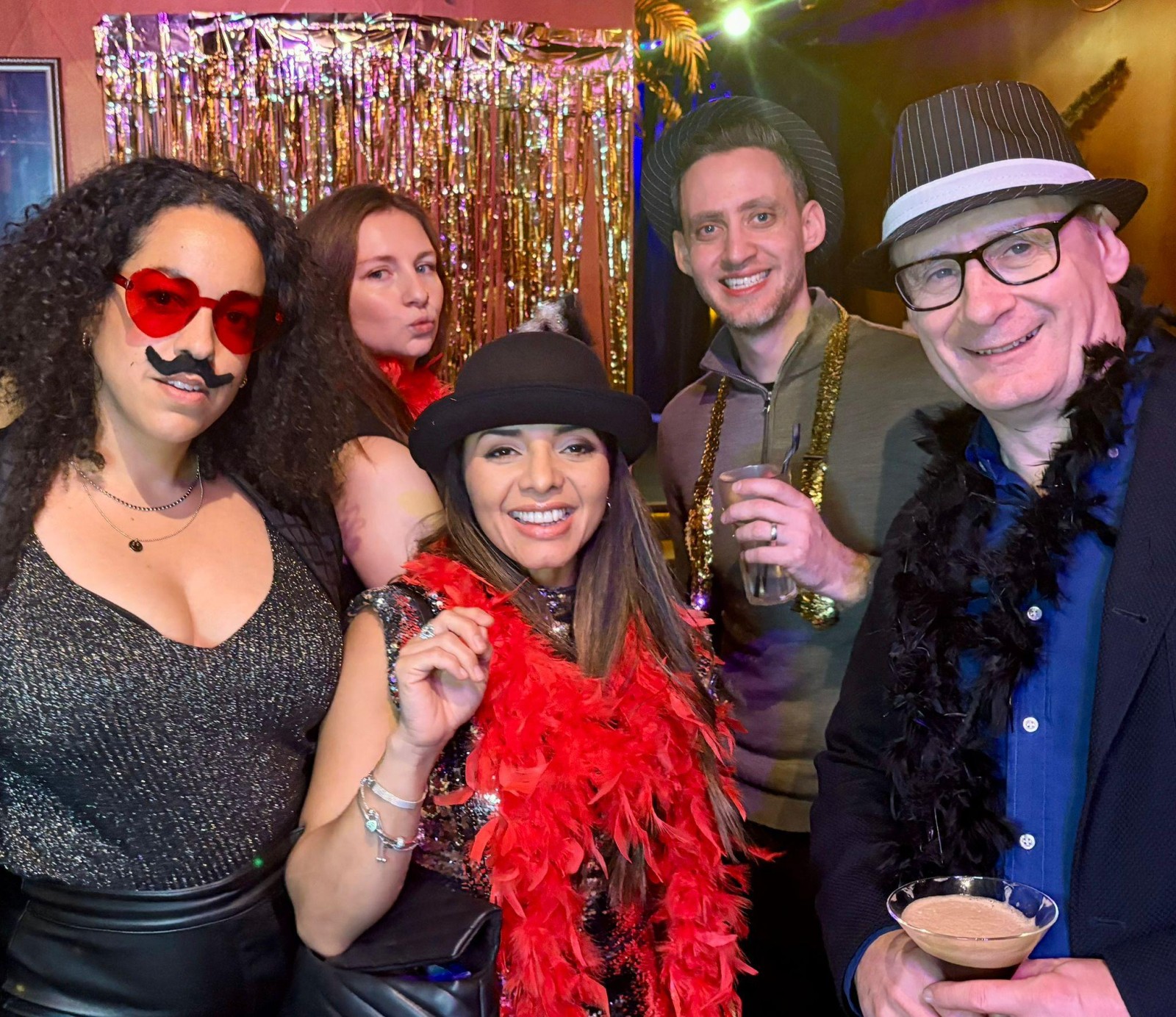 A lively group of five friends pose for the camera in a festive setting, adorned with shimmering gold and red decorations. Clad in black and vibrant outfits, they wear playful accessories, including feather boas and quirky glasses. The atmosphere exudes celebration.