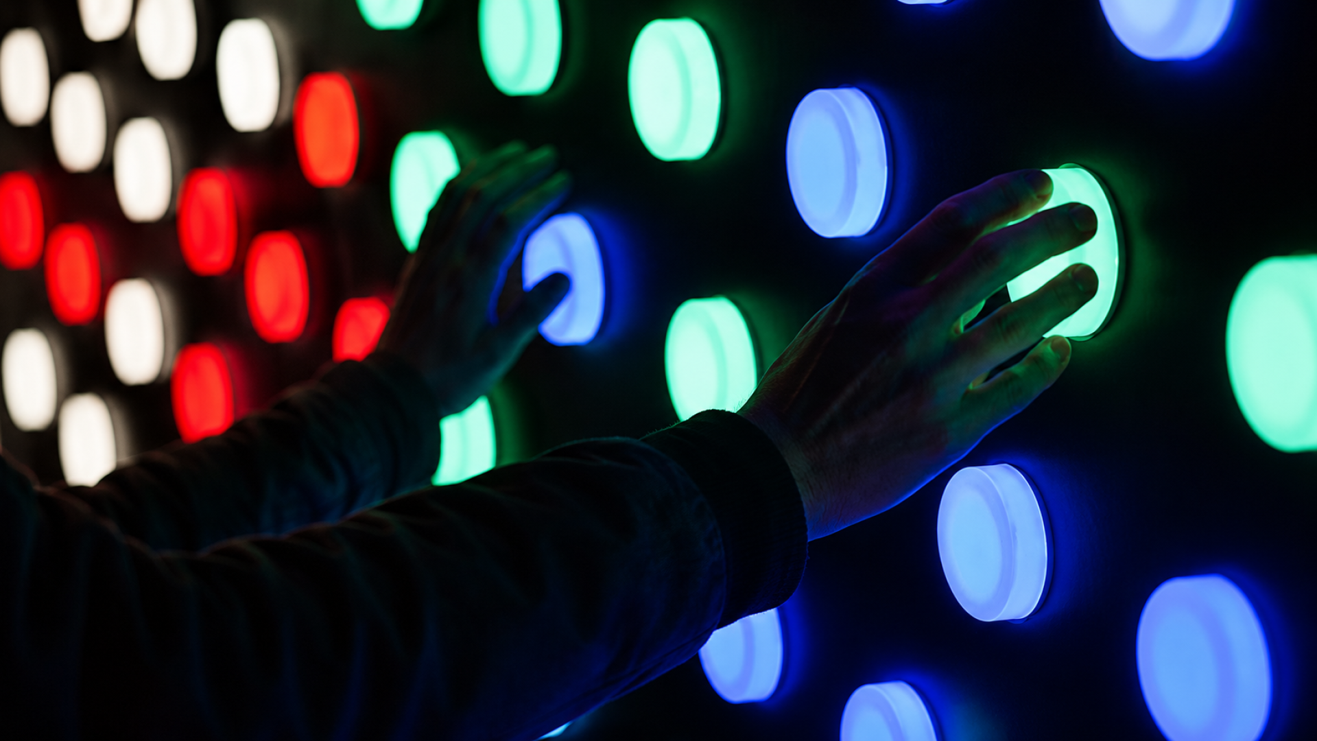 Two hands hover over a vibrant, illuminated panel featuring an array of circular buttons, glowing in red, green, blue, and white. The dark background creates a striking contrast, enhancing the vivid colours and inviting interaction with their smooth, textured surfaces.