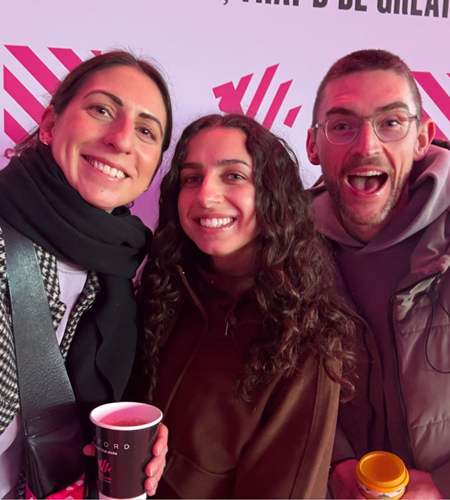 A cheerful trio stands close together against a vibrant backdrop adorned with striking pink geometric patterns. Each person holds warm cups, wearing stylish attire suitable for a casual outing. The warm lighting enhances their relaxed atmosphere, highlighting the textures of their clothing.