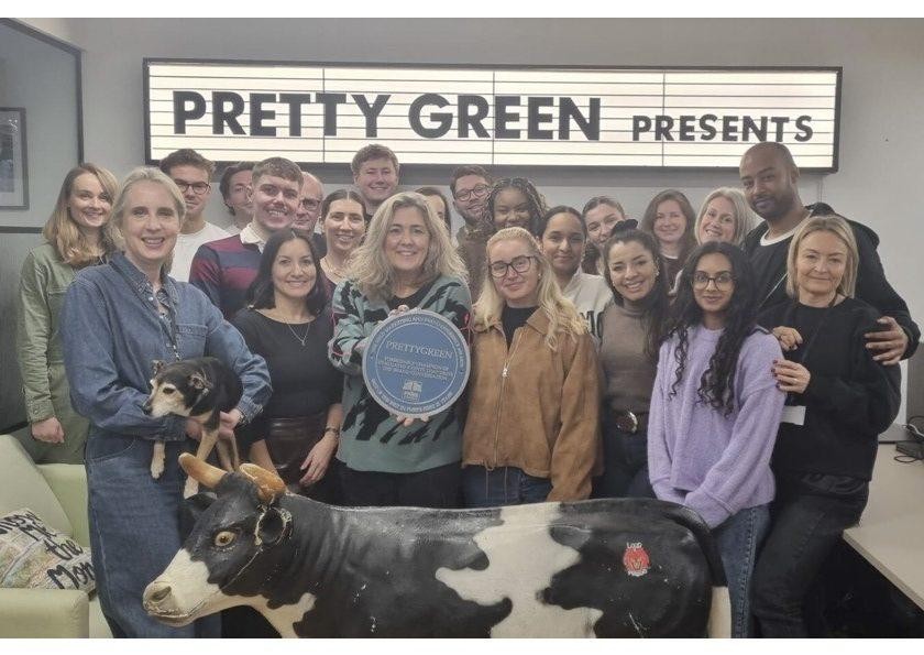 A vibrant group of individuals stands together in an office space, with a large lightbox displaying 'Pretty Green Presents' behind them. Various personalities, including a woman holding a small dog and another showcasing a round plaque, gather around a life-sized black and white cow sculpture. The atmosphere is cheerful and collaborative, reflecting a creative environment.