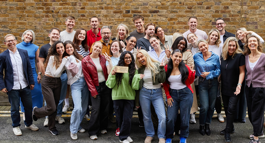 A vibrant group photo featuring a diverse assembly of individuals, standing together against a textured brick wall. Some hold a wooden plaque, while others strike playful poses. Their outfits range from casual to smart, showcasing an array of styles and colours, embodying a lively, collaborative atmosphere.