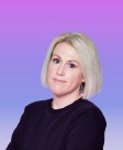 A woman with shoulder-length, straight platinum blonde hair stands against a smooth gradient background transitioning from pastel purple to light blue. Wearing a black sweater, she gazes slightly off to the side, conveying a contemporary aesthetic characterized by simplicity and elegance.