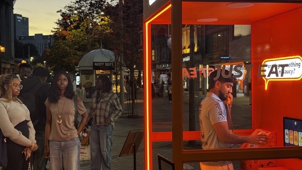 A vibrant neon-orange food kiosk stands on a bustling city street at dusk. A young man in a grey shirt speaks on a phone inside the booth, illuminated by warm light. Nearby, a small group of people observes the scene, framed by subtle autumn foliage and urban architecture.