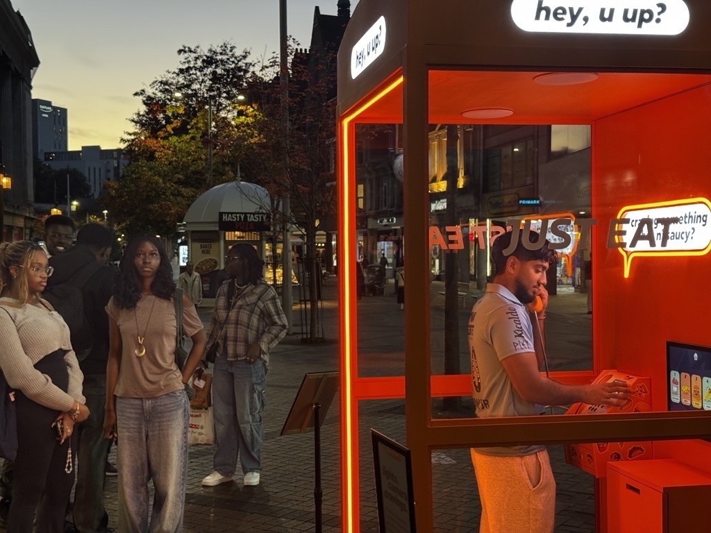 A vibrant orange food kiosk glows in the evening light, showcasing bold signage that reads "Hey, u up?" A young man in a grey outfit interacts with a digital menu inside the booth. Nearby, a group of people watch curiously, framed by the bustling urban backdrop.