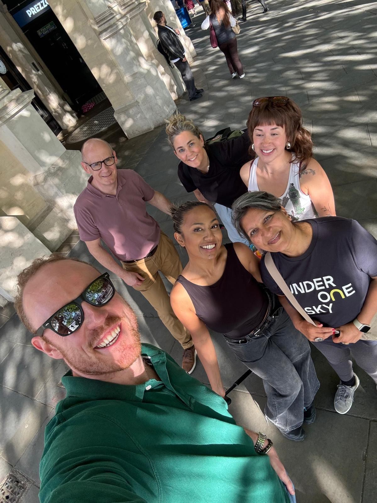 A group of five people posing for a cheerful selfie in a bustling urban area. The sun filters through trees, casting playful shadows on the pavement. They are casually dressed, exuding a relaxed vibe, with a lively backdrop of pedestrians and buildings.