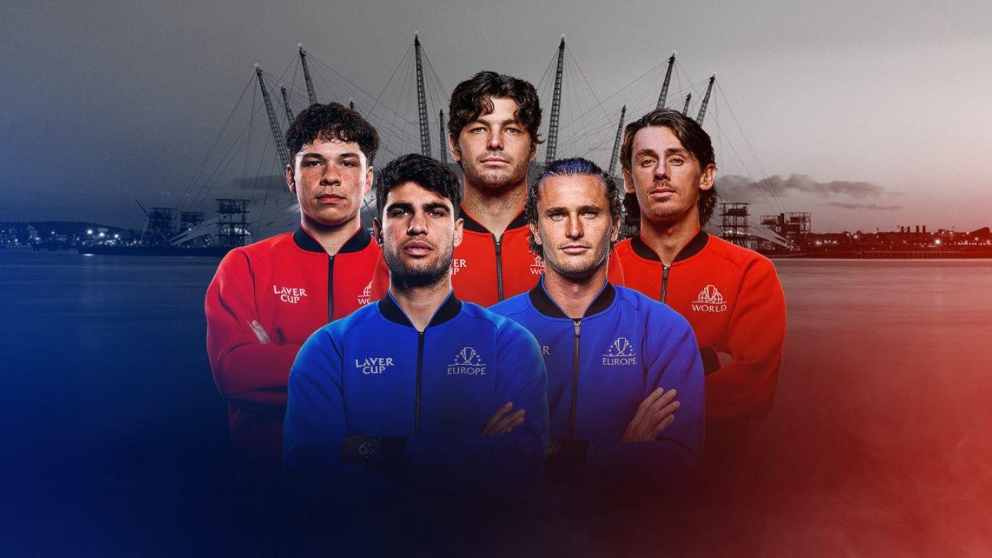 A striking group portrait of five individuals standing confidently in front of a blurred waterfront backdrop. Each person wears a distinctive coloured jacket—red, blue, or black—adding to the dynamic composition. The setting features a dramatic skyline with cranes in silhouette, enhancing the industrial atmosphere.