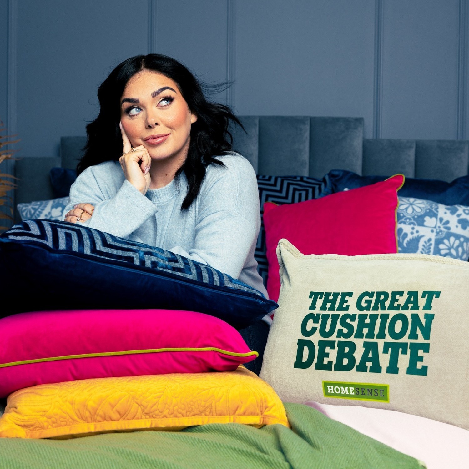 A woman in a light grey sweater leans thoughtfully on a stack of vibrant cushions, including a layered pink and bright yellow piece. Behind her, a textured blue wall complements the luxurious upholstery of the bed, creating an inviting atmosphere for the 'Great Cushion Debate' theme.