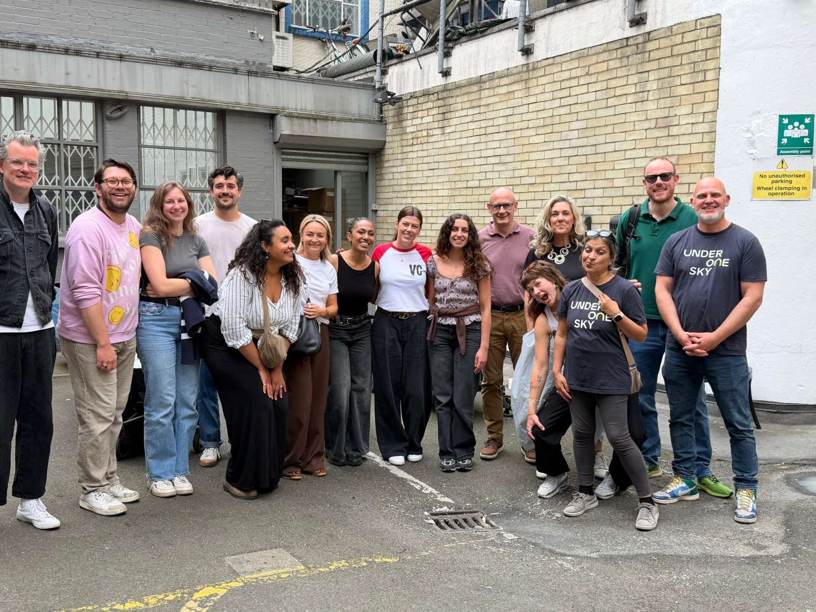 A diverse group of ten individuals poses together in a narrow, bustling alley. They wear casual clothing, with a mix of bright and muted tones. The textured stone wall provides a contrasting backdrop. Sunlight highlights their relaxed stances, creating a vibrant atmosphere.